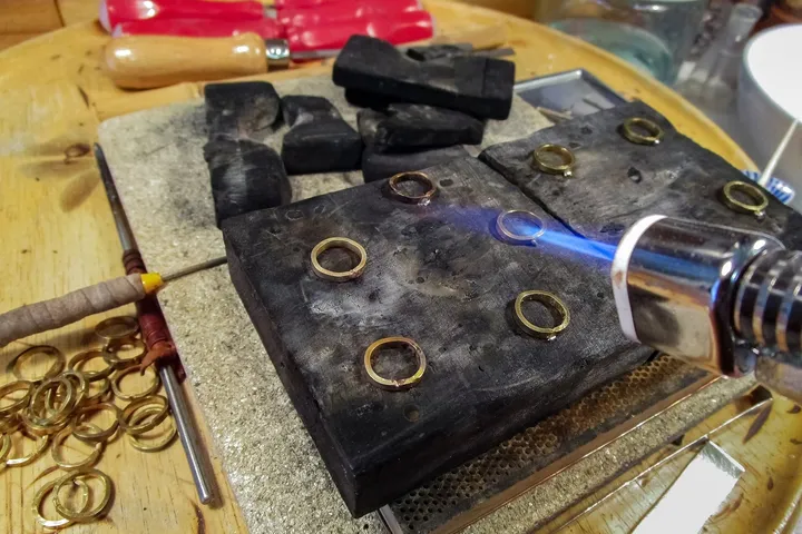 A gas torch playing over brass rings laid out on a soldering block, with a ring mandrel and hand tools beside them