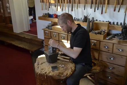A silversmith at a workshop bench with hammers and stakes behind him